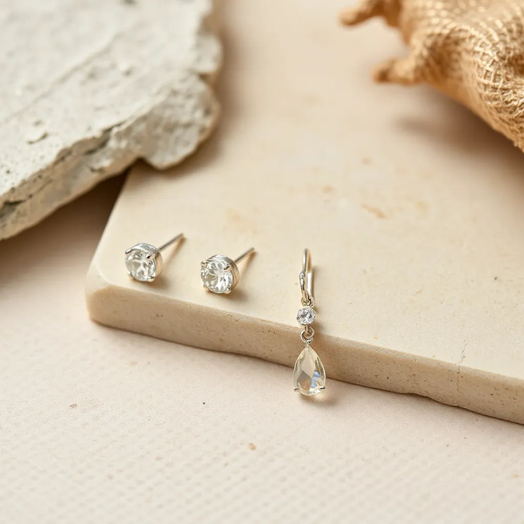 Elegant silver earrings in a minimalist close-up, showing a stud and a delicate drop design on a neutral surface.